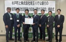 杉戸町窪田裕之町長と株式会社埼玉りそな銀行平野隆司執行役員埼玉東地域営業本部長が両者関係者他５名と、協定式で協定締結を記念して協定書を掲げる様子
