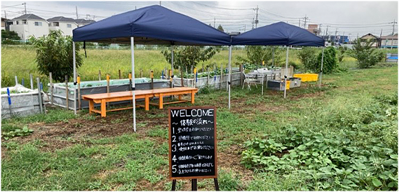 畑の中に青いテントとベンチが設置され、手前に「WELCOME」と書かれた案内板が立っている屋外の様子