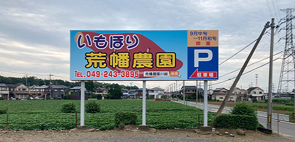 畑の前に「いもほり 荒幡農園」と駐車場案内が書かれた看板が立っている写真