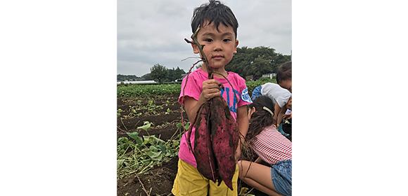 畑で、ピンクの服を着た子どもが土の付いた芋を持って立っている。周囲にほかの子どもがいる。