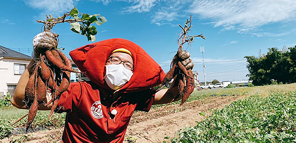 畑でマスクを着けた人が、掘り出したサツマイモを両手に持っている写真