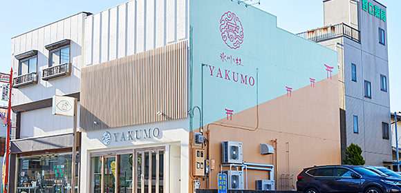 「YAKUMO」と書かれた店舗外観。水色と薄いオレンジ色の壁にピンクの鳥居の模様があり、右側に車が停まっている。