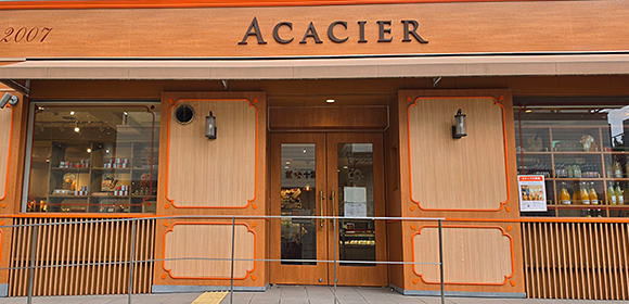 「ACACIER」と書かれた看板のある店舗外観。木目調とオレンジ色の装飾の正面に、ガラス扉とショーウィンドウがある。