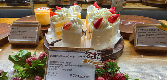 ショーケース内に、イチゴがのったショートケーキやプリンが木製台に並び、商品名と価格の札が置かれている。