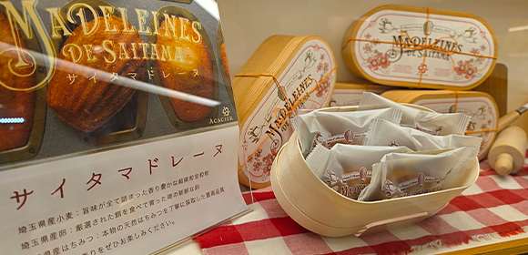 「サイタマドレーヌ」と書かれた焼き菓子のパッケージと、個包装の商品が並ぶ店頭ディスプレイ。