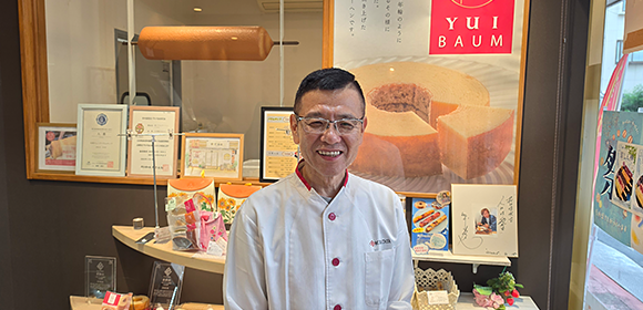 白い調理服を着た男性が、バウムクーヘンのポスターや商品が並ぶ店頭の前で立っている。